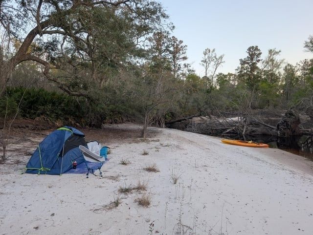 Tent and folding chair on a sandy riverside campsite beside a kayak