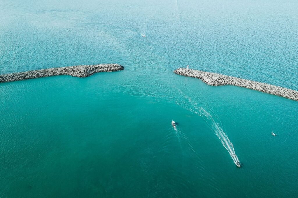 Boats pass between two concrete breakwaters in turquoise water